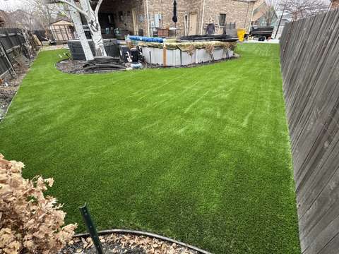 A fenced backyard lawn of artificial turf behind a brick house, with patio furniture
