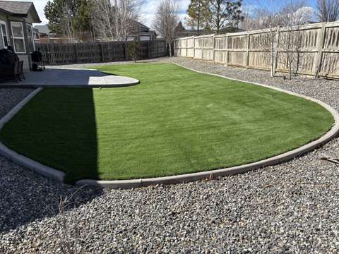 A curved artificial turf lawn inside a concrete border, gravel beds and a wood fence around it