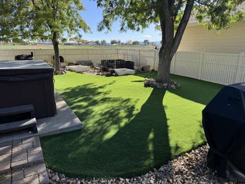 An artificial turf lawn around a hot tub and a large shade tree, with a vinyl fence behind