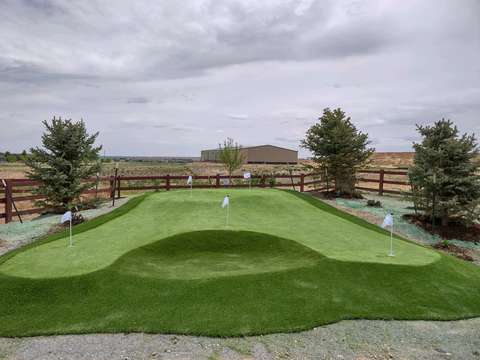 A backyard putting green shaped with a raised contour and three flags, a split-rail fence and plains beyond