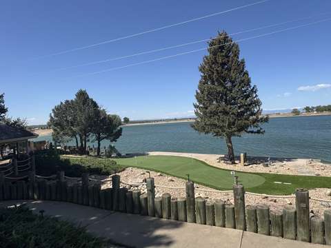 A putting green on a lakeside lot behind a timber fence, with a pine tree and the water beyond