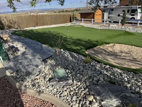 A backyard putting green with a sand bunker, river rock beds and a flagstone path, open plains beyond