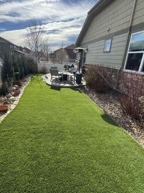 A narrow side-yard lawn of artificial turf between a house and a planting bed, leading to a patio