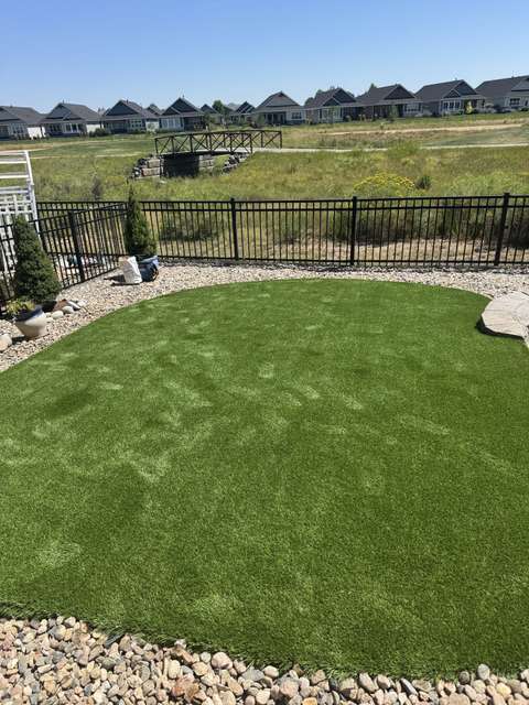 An artificial turf back lawn inside a black iron fence with a river-rock border, open space beyond, near Windsor, Colorado