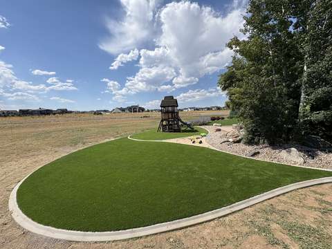 A curving artificial turf lawn edged by a concrete curb, with a wooden playset and river rock, near Windsor, Colorado