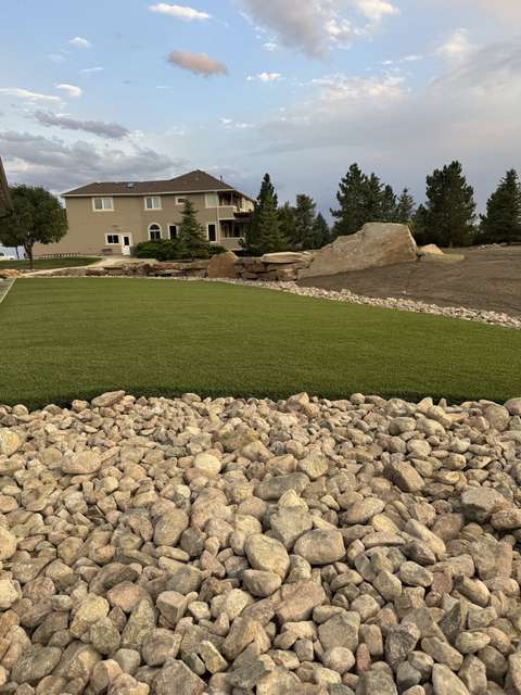 A new artificial turf lawn edged with river rock below a two-story house and boulder wall, near Berthoud, Colorado
